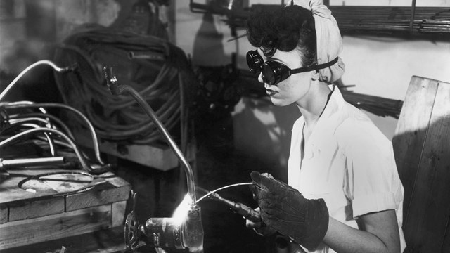 A black and white image of a woman in goggles and gloves welding.  