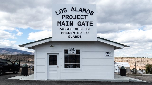 A small white building with a sign that reads "Los Alamos Project Main Gate"