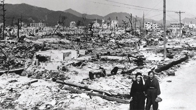 A black and white photo of destroyed buildings as two people walk along the road. 