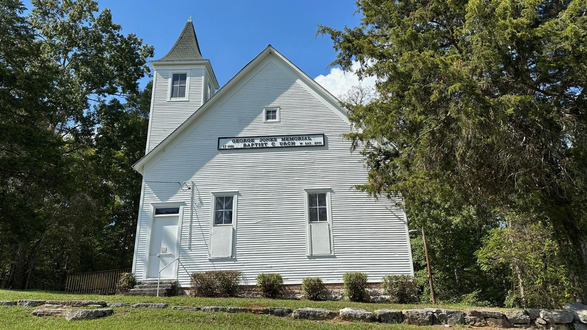  A white wooden church on a grassy hill surrounded by trees.