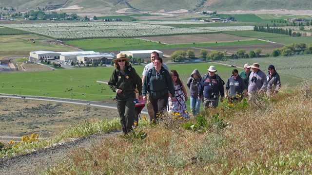 7 adults and 1 kid hike behind a uniformed ranger up the side of a grassy hill on a clear day.