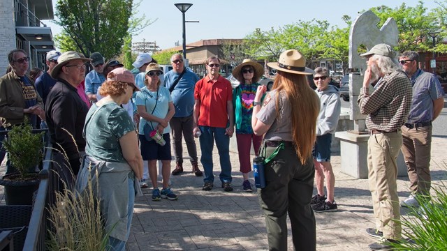 15 adults stand on the sidewalk in a semicircle around a uniformed ranger on a sunny day.