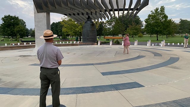 Park ranger faces a large bronze bell overhanging a courtyard about to be rung by a wooden striker.