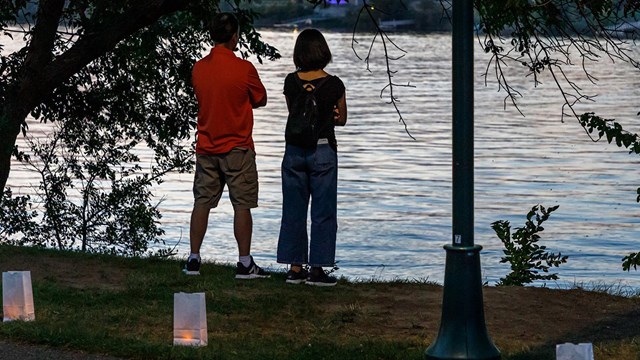 Two people look out on a river with luminarias behind them. 