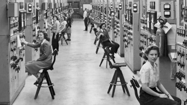 Several women seated at stools along a long hallway with equipment along the walls.