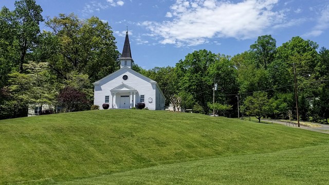 A green grassy hill with a white chapel on the top and a historic sign in the foreground.