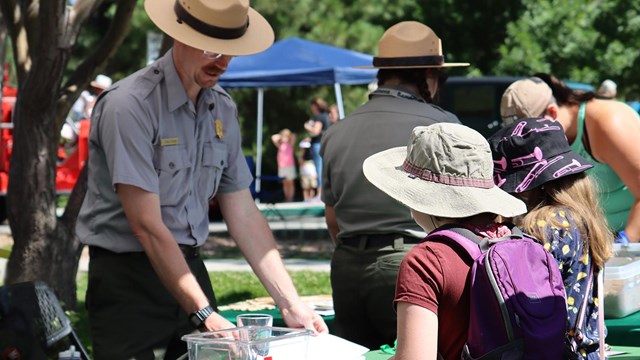 A man in a ranger hat shows children a piece of paper on the table.