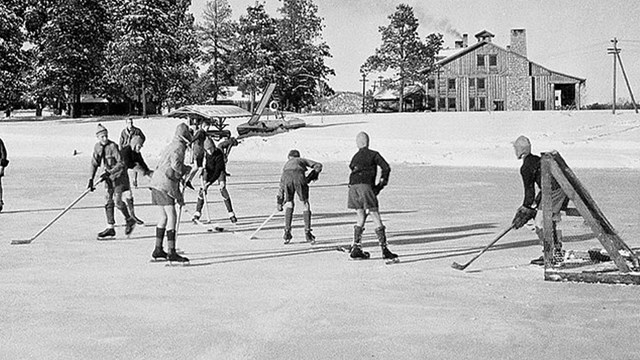 Several youths play ice hockey on a frozen pond with a house and trees in the background.     
