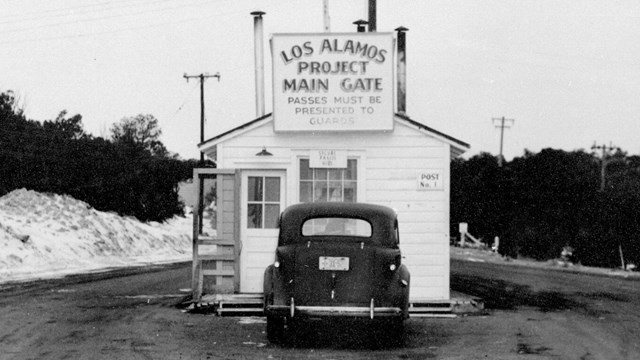 A small white sided building with a sign reading "Los Alamos Project Main Gate" on a muddy road.