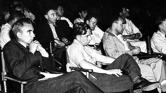Men sit facing an unseen presenter in rows of folding armchairs atop a scuffed wood floor.