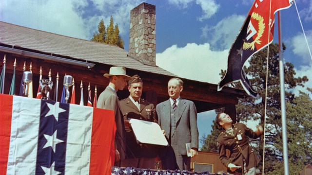 A color of three men standing on a stage in front of a log building. 