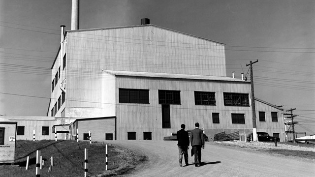 A black and white photo of a large white building with two people walking toward it.  