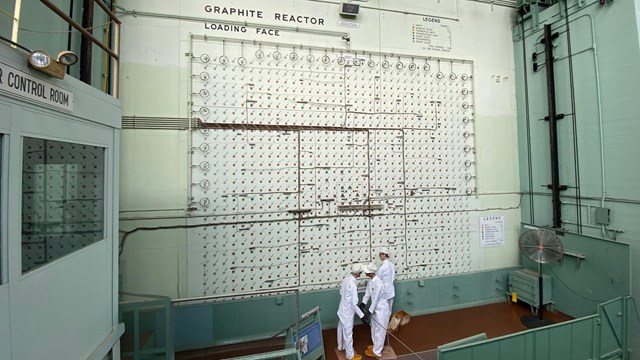 Three mannequins in white clothes stand at the base of a large reactor face. 