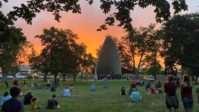 Several dozen people gather in a city park at sunset.