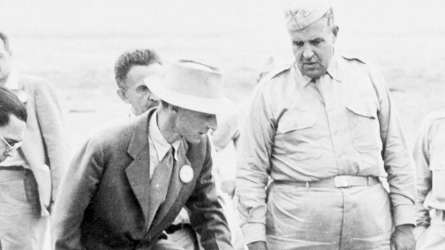 Two men stand in the desert looking at the ground directly in front of them.