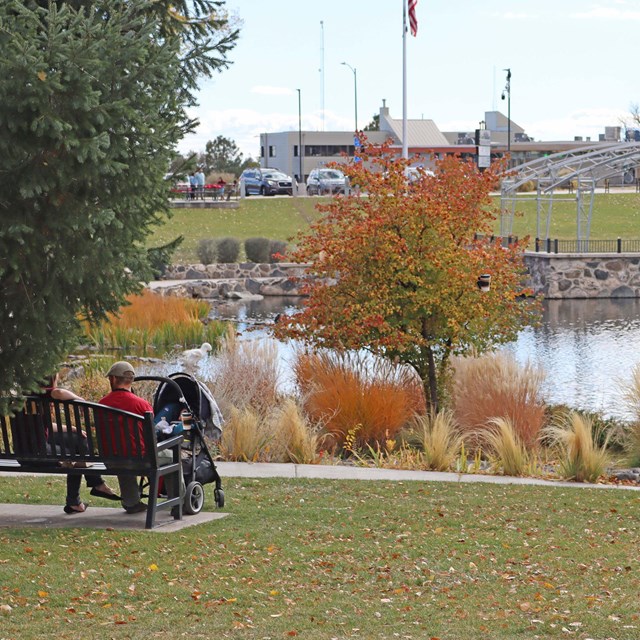 A couple with a stroller sit in front of a lake surrounded by fall colors.