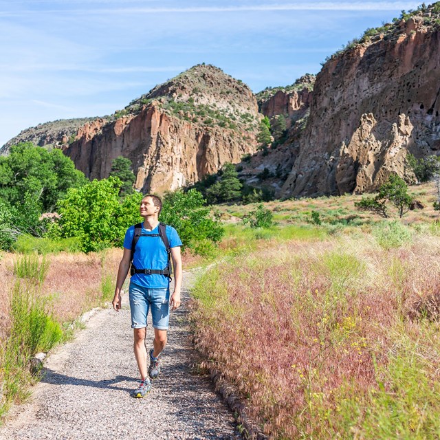 A man in a short-sleeved shirt and shorts hikes along a gravel path in a desert with scrubby vegetat