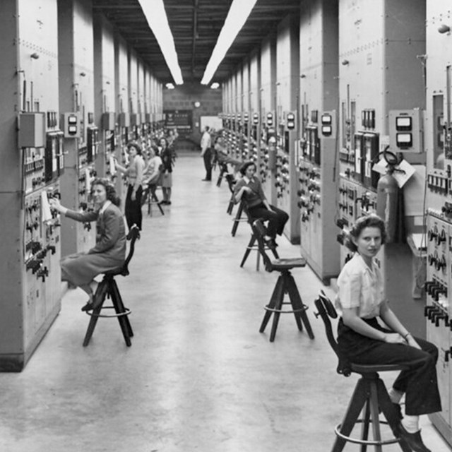 Several women seated at stools along a long hallway with equipment along the walls.