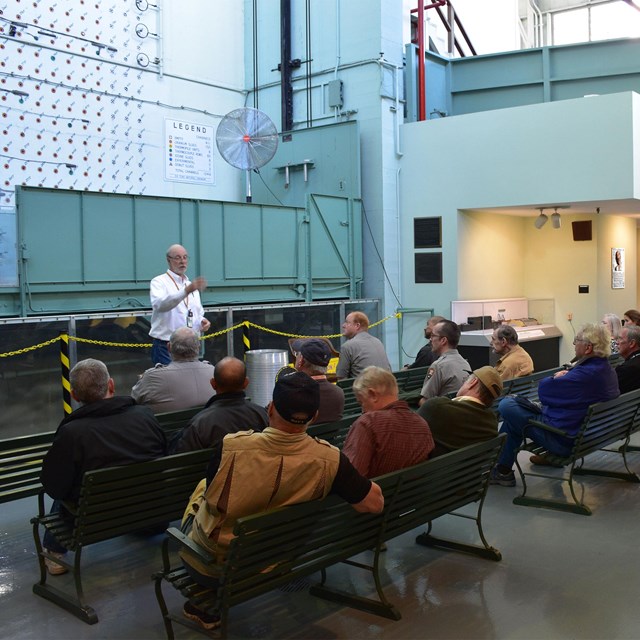 Several people seated on benches watching a speaker in front of a reactor face. 