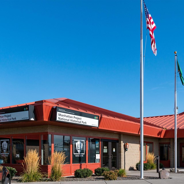  Exterior photo of a one-story visitor center with a red roof and two flag poles in front of the bui