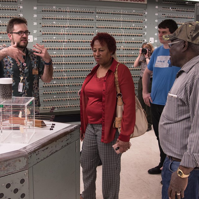 A group of people stand around a man who is gesturing. There is a table and silver background. They 