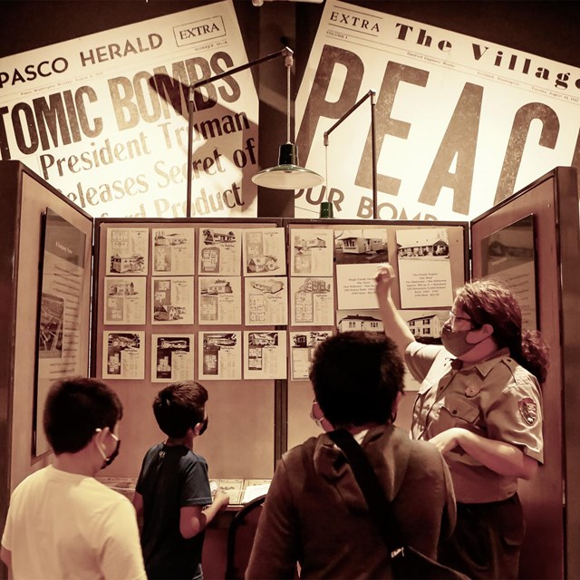 A woman in a uniform is pointing to pieces of paper displayed in a cubicle to three children. On the