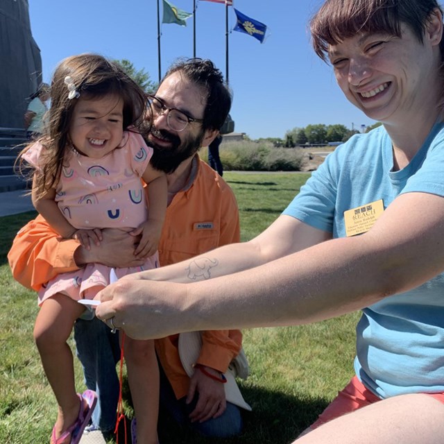 A smiling man holds a smiling child while a young woman holds onto a paper crane with her arms outst