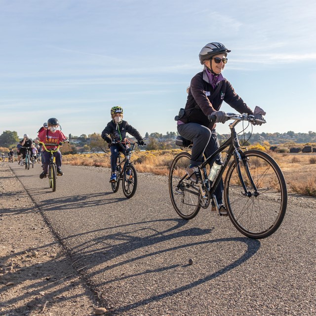  A woman and two young boys bicycle down a paved path next to bushes. The sky is blue