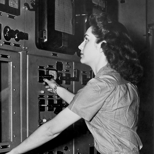 A black and white photo of a woman in front of a wall of dials.