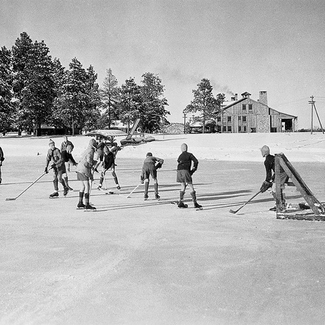 Several youths play ice hockey on a frozen pond with a house and trees in the background.