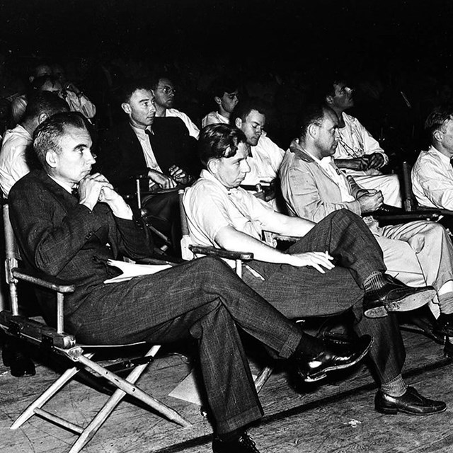 Men sit facing an unseen presenter in rows of folding armchairs atop a scuffed wood floor.