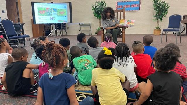 A ranger seated in a rocking chair reads a book to a group of children gathered around her 