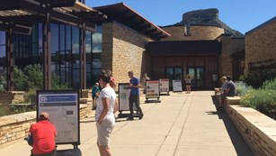 People study signs along a walkway in front of a large stone visitor center building