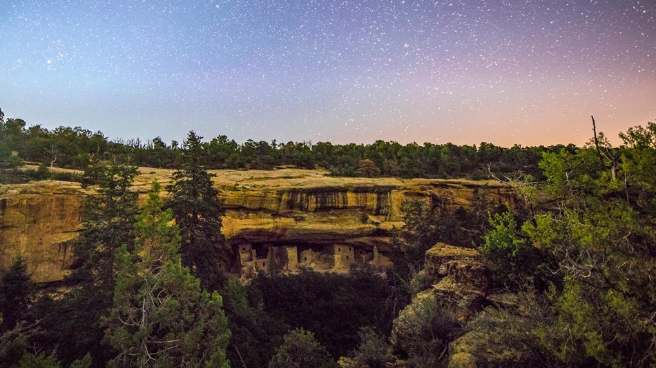 The moon rises into a dark sky full of stars above a sandstone cliff containing an ancestral village