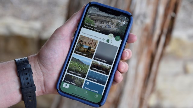 A hand holds a phone with the National Park Sercice app displayed on the screen in front of a tree