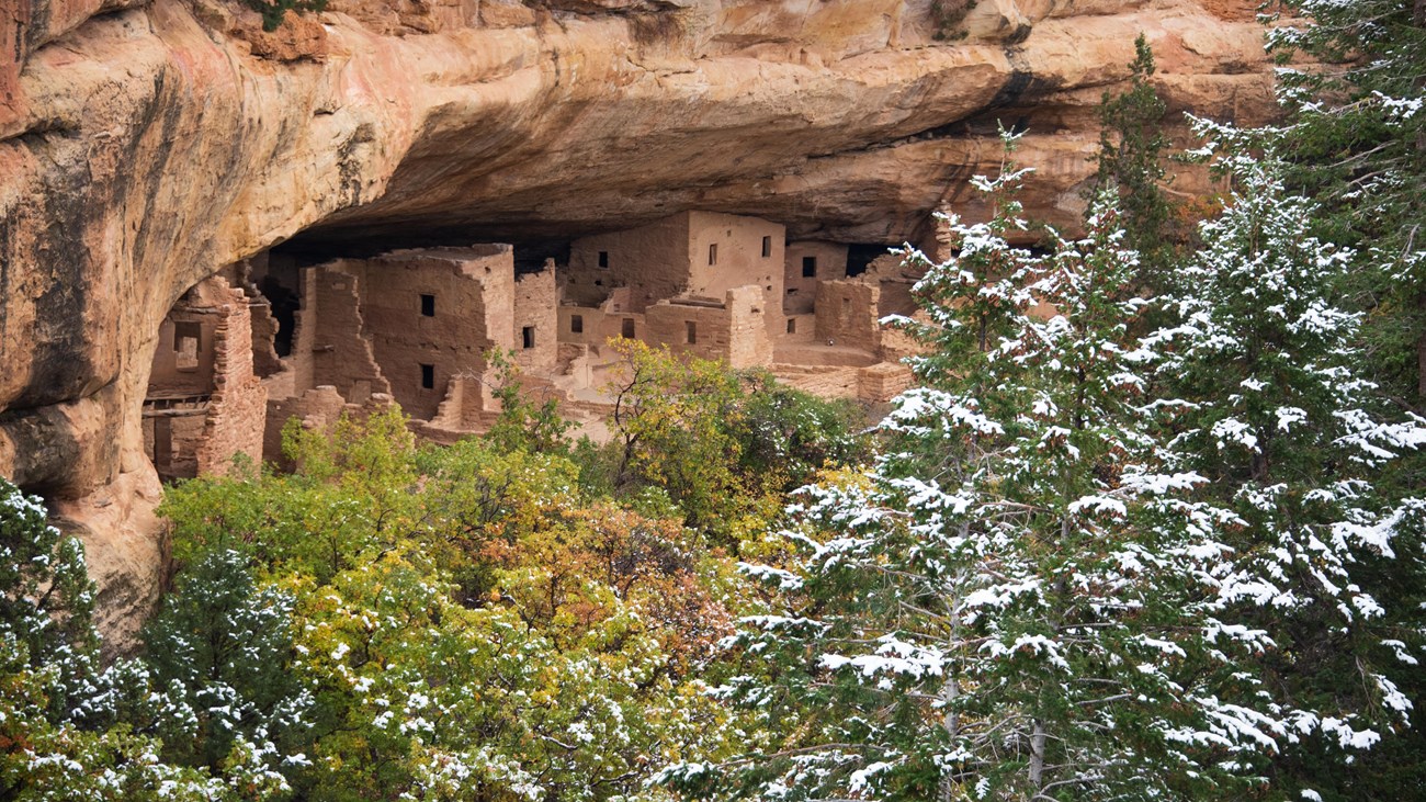 A stone masonry village under a sandstone alcove and behind snow-covered firs