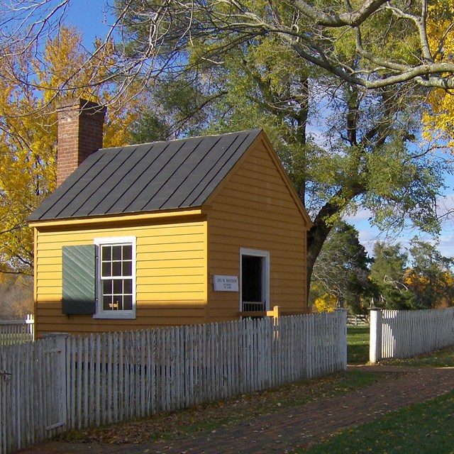 Small yellow A-frame building behind a white fence, surrounded by trees turning from green to yellow