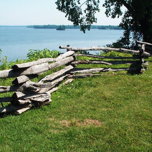 A stacked log fence at the edge of a field, with a view of a vast river beyond