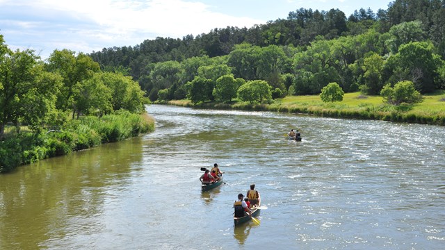 Bird's eye view of three canoes on a river under blue sky. Green forest and prairie lines the river.