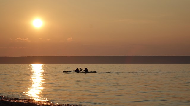 Two people in a tandem kayak paddling while the sun is setting on the horizon.