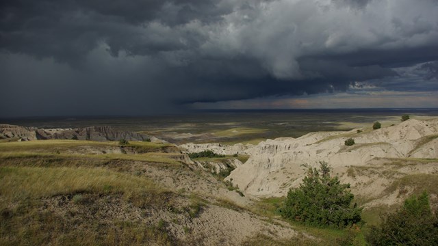 Dark clouds over prairie and a white-colored canyon with a few small trees. 