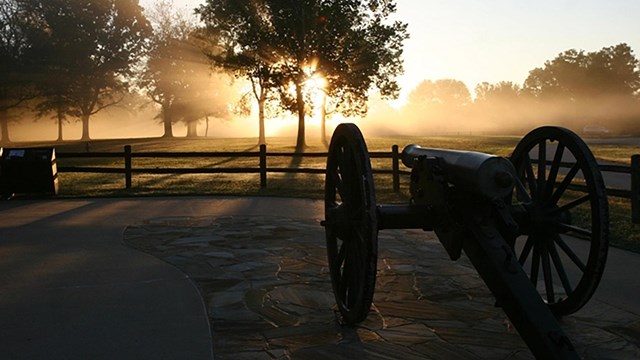 Rays of light from sunrise shine past a cannon sitting in a courtyard. 