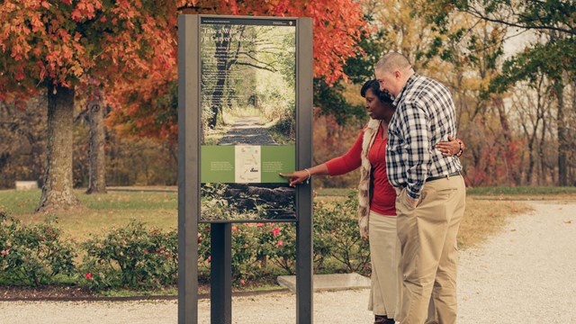 A man and woman looking at an wayside exhibit along a walking trail.