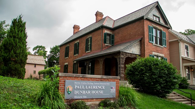 A sign stands out front of a brick house on an overcast day. 