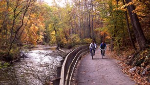 Two bicyclists on a path by a river during fall. 