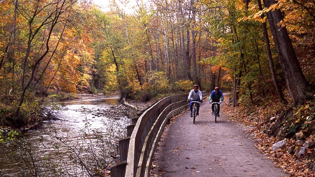 Two bicyclists on a path by a river during fall.