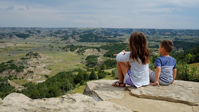 Two children sit on the edge of a rock ledge and gaze out over the badlands, juniper, and trails.