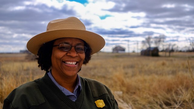 Park Ranger from chest up standing in front of a brown field with a schoolhouse in the distance.
