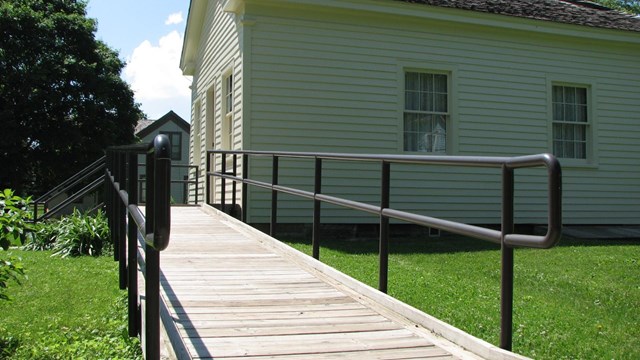 A wooden ramp with a metal railing leads to the front door of an old schoolhouse.