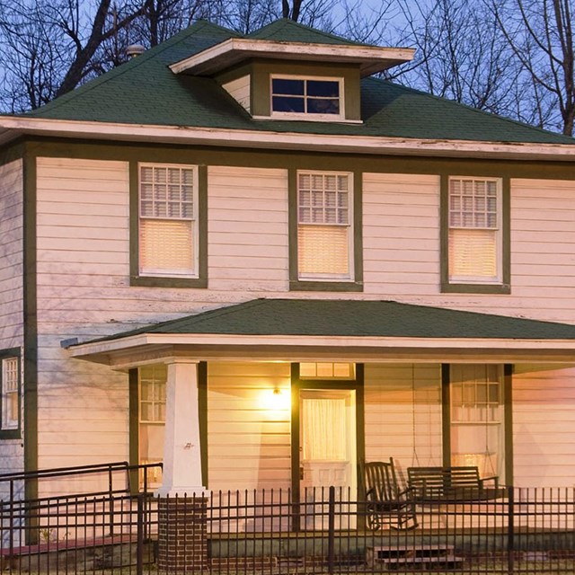 Two story white home with green trim lit at dusk in front of trees. 